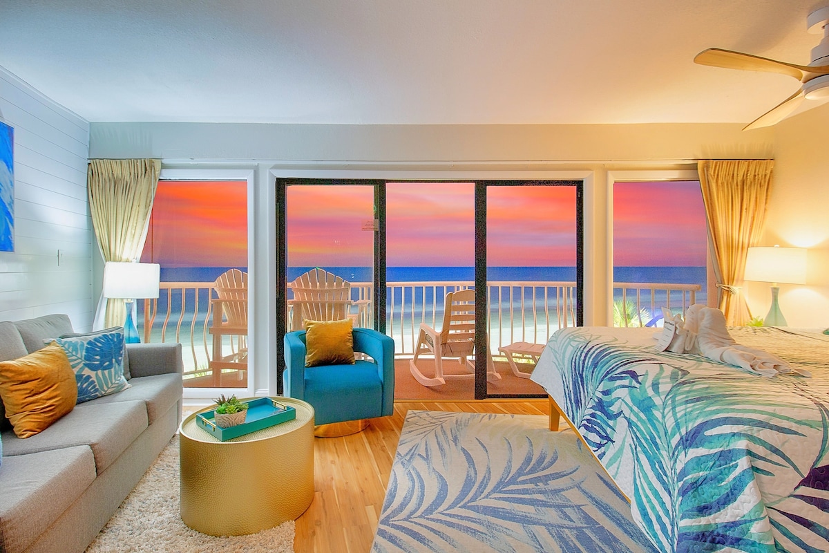 A spacious living area features a king bed with vibrant bedding, a comfortable gray sofa, and a blue armchair. Large glass doors reveal a stunning view of the ocean at sunset, creating a serene ambiance. The room is illuminated by soft lighting, enhancing its relaxing atmosphere.