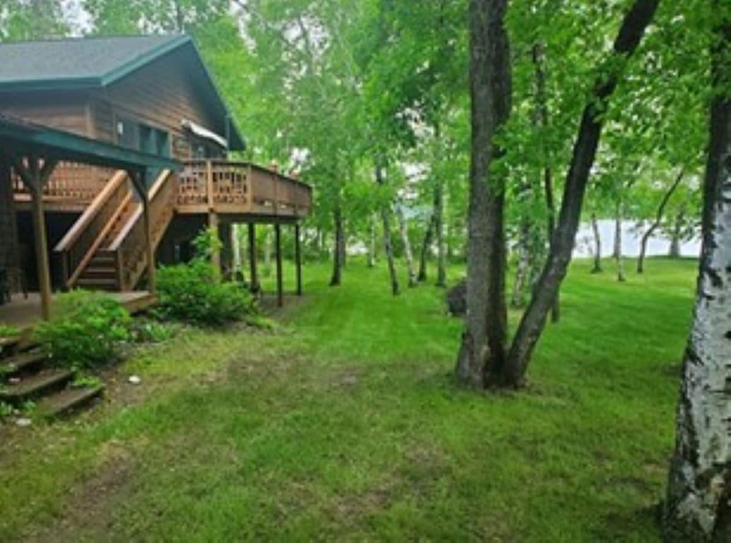 The cabin is set among lush greenery, with a spacious deck overlooking a manicured lawn. Steps lead from the yard to the deck, which is framed by trees. The lake can be seen in the distance, enhancing the natural surroundings.