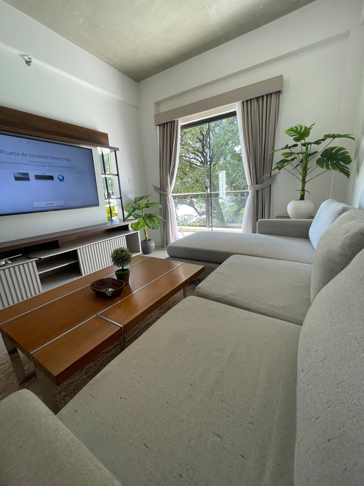 A light-filled living area features a modern sectional sofa in neutral tones, complemented by a wooden coffee table. A large television is mounted on a sleek entertainment unit. Natural greenery is visible through the nearby window, offering a view of the tranquil outdoor space.