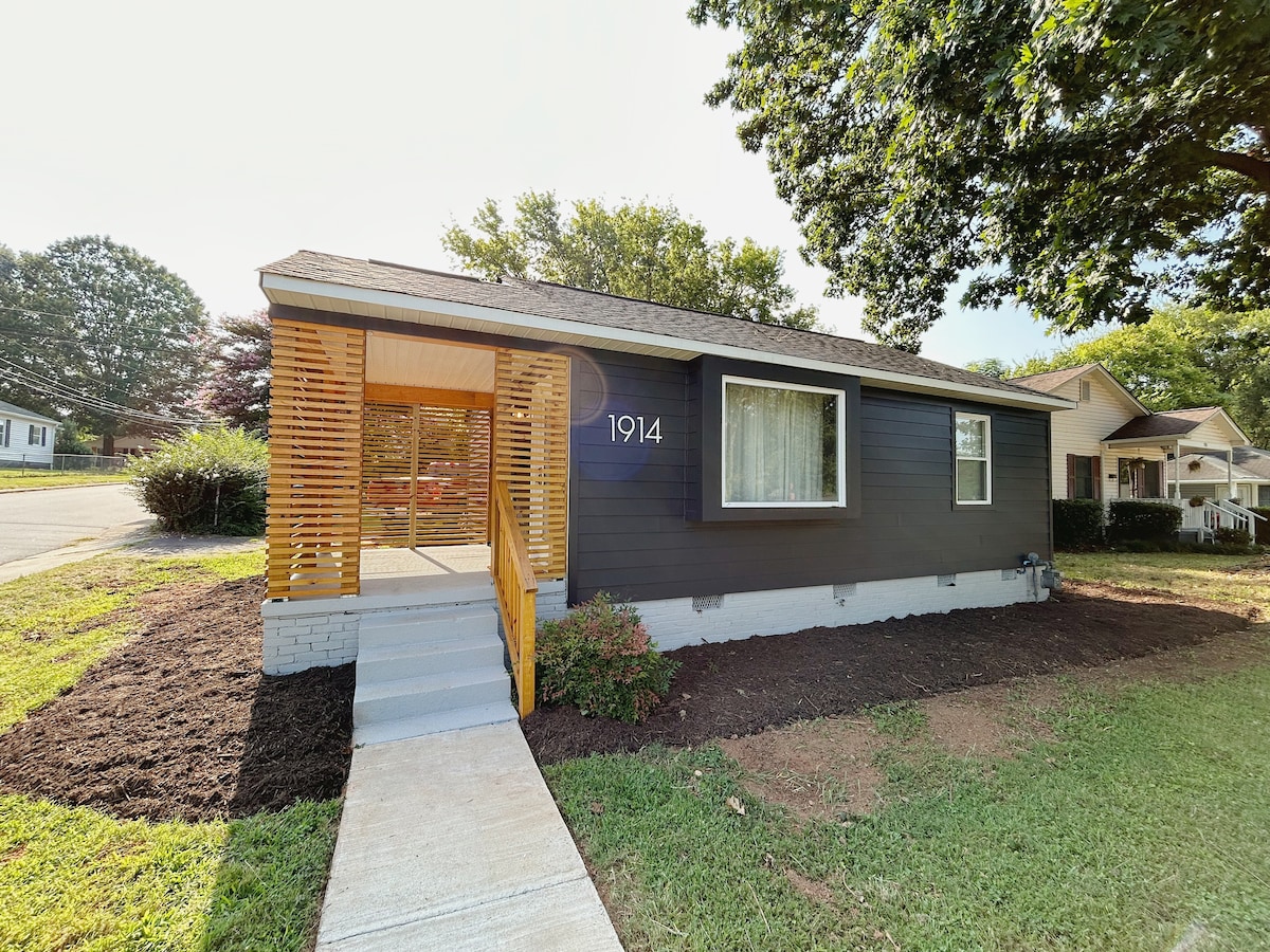 The exterior of the home features a modern design with a dark-colored facade and light wooden accents. A pathway leads to the front porch, where large windows allow natural light into the living area. Neat landscaping surrounds the entrance, creating a welcoming approach.