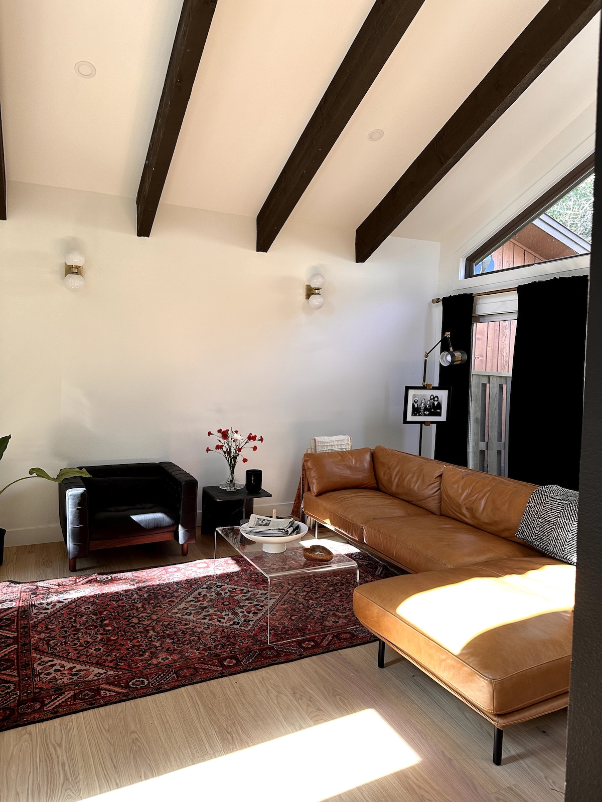 A light-filled living room features vaulted ceilings with exposed beams. A brown sectional sofa is positioned near a decorative area rug. A small black chair and coffee table are also present, complemented by plants and wall art. Large windows provide natural light.