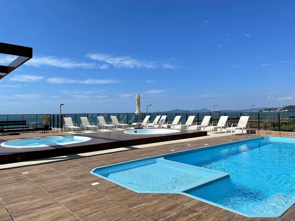A spacious outdoor leisure area features multiple pools, including a large main pool and Jacuzzis. Sun loungers are arranged around the pool deck, with a view of the ocean in the background. Clear blue skies enhance the inviting atmosphere of the space.