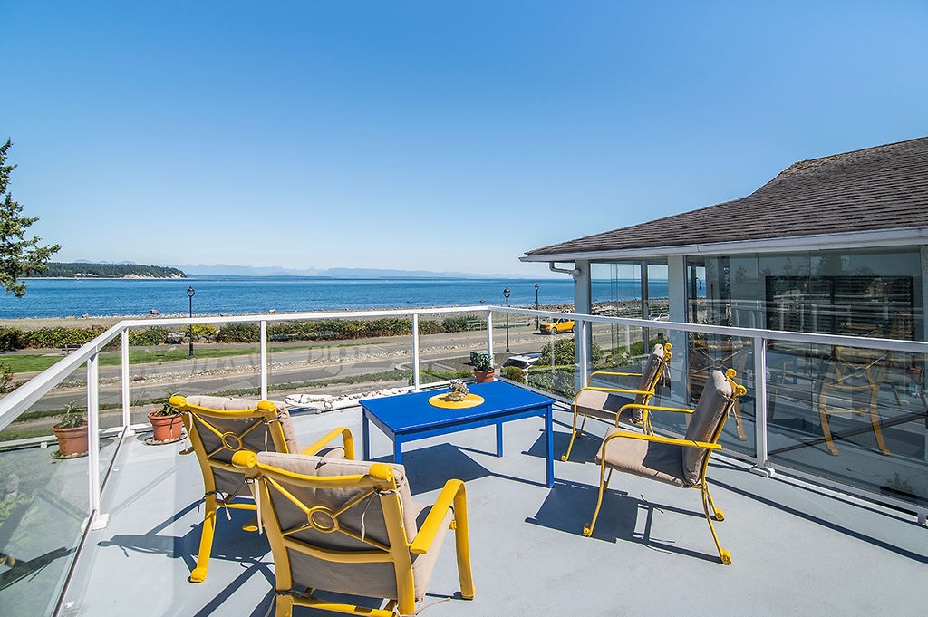 A spacious deck showcases bright yellow outdoor chairs surrounding a blue table. The scene offers unobstructed views of the Salish Sea and distant mountains, with greenery and the Seawalk visible below. Sunlight enhances the inviting atmosphere of this outdoor space.