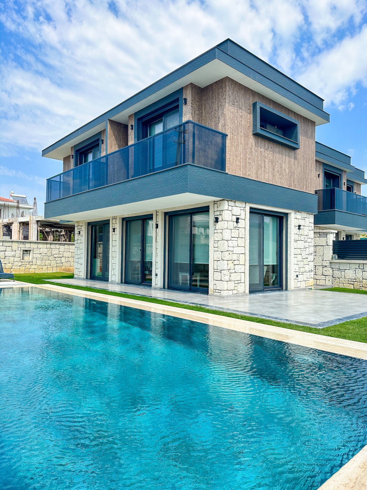 A two-story villa is shown, constructed with a blend of stone and modern materials. Expansive glass doors lead to a spacious patio, offering views of a clear blue pool. A balcony extends from the upper level, providing additional outdoor space.