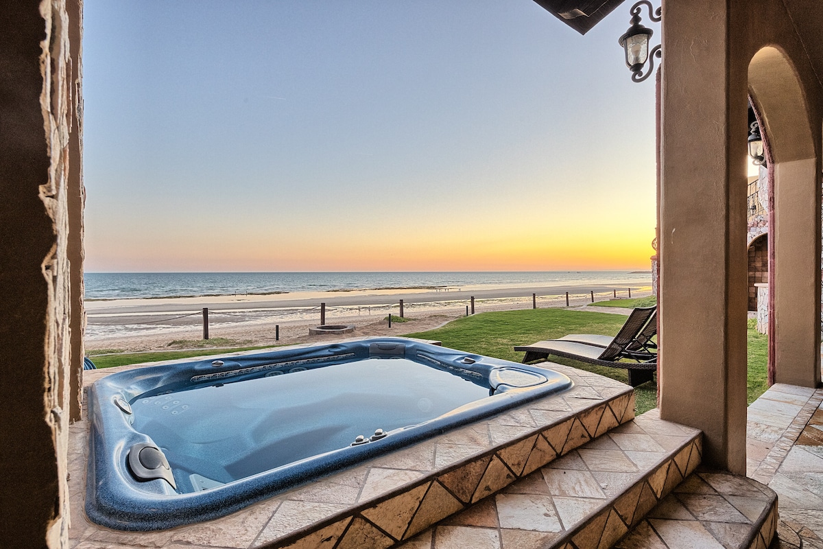A private jacuzzi is positioned on a patio, offering a direct view of the ocean at sunset. The tranquil scene features gentle waves and a gradient sky transitioning from orange to deep blue. Grass surrounds the jacuzzi, and lounge chairs are neatly arranged nearby.