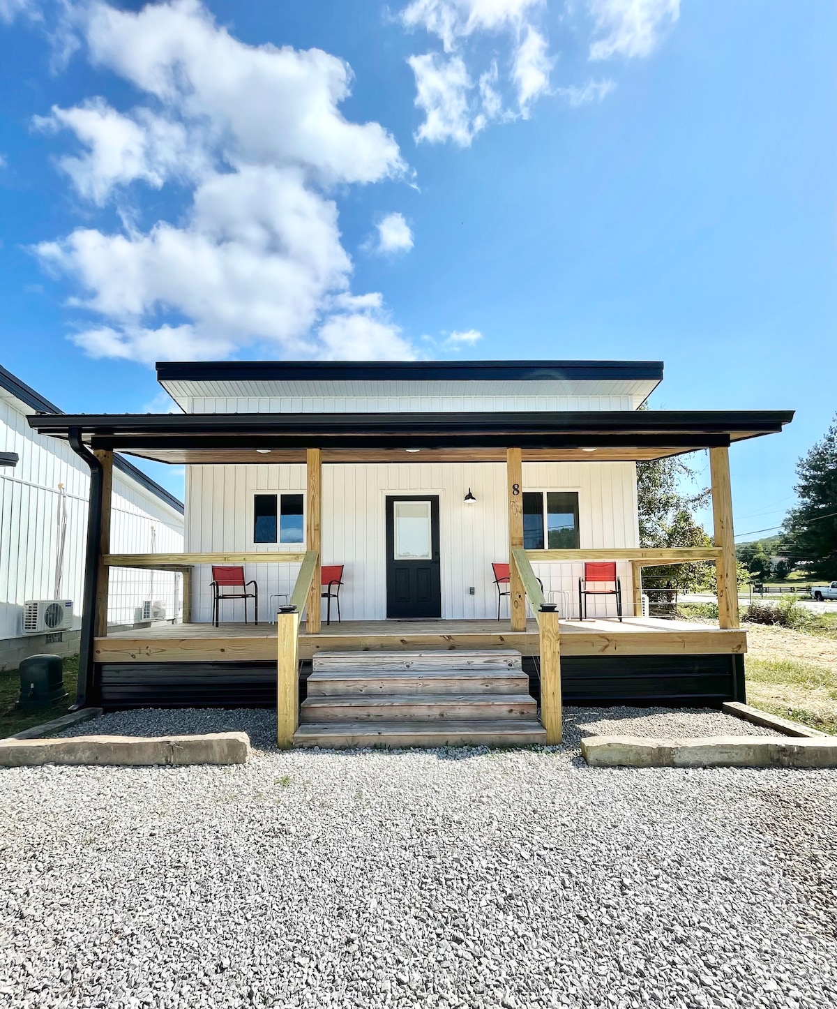 A modern tiny home features a welcoming front porch with two red chairs positioned beside a small table. Steps lead up to the entrance, which is framed by a simple black door. The building's exterior showcases a white facade with large windows allowing natural light.