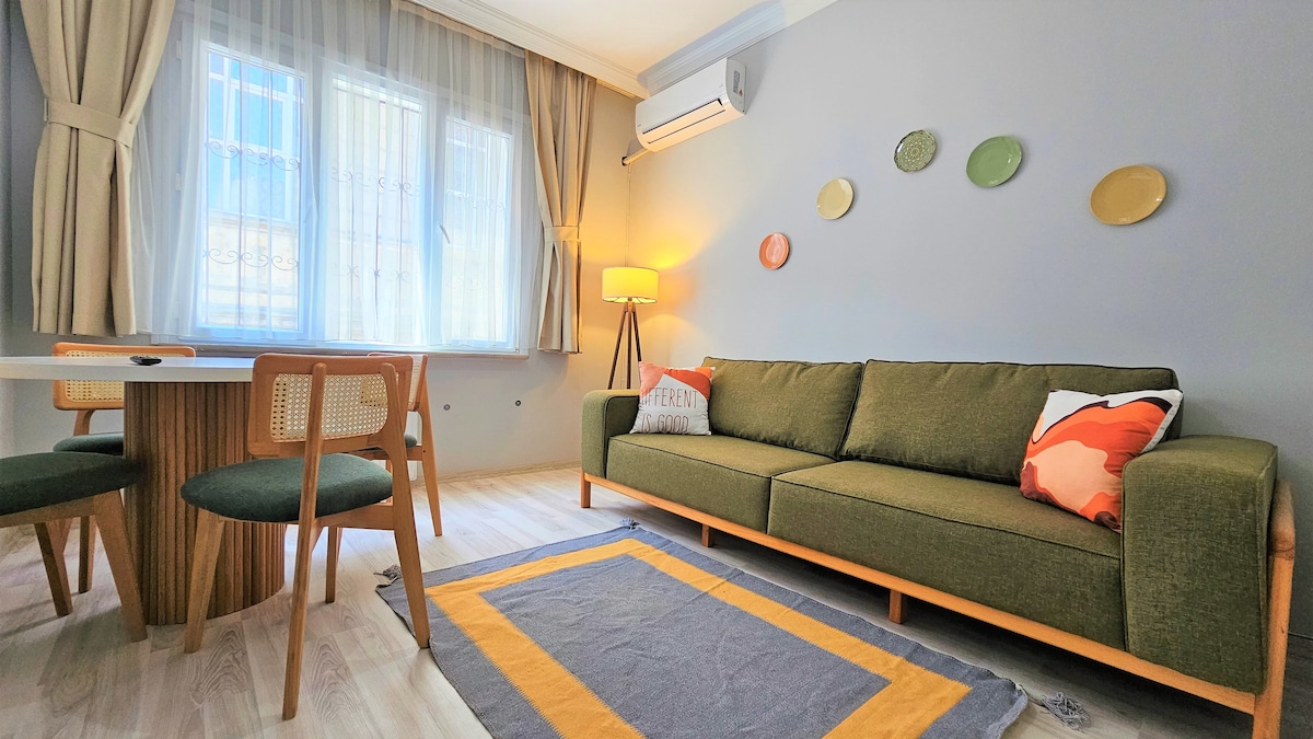 A living area features a comfortable green sofa with accent pillows, complemented by a wooden dining table and chairs. Decorative plates are arranged on the wall, and natural light fills the space through sheer curtains. A patterned rug adorns the floor, enhancing the atmosphere.