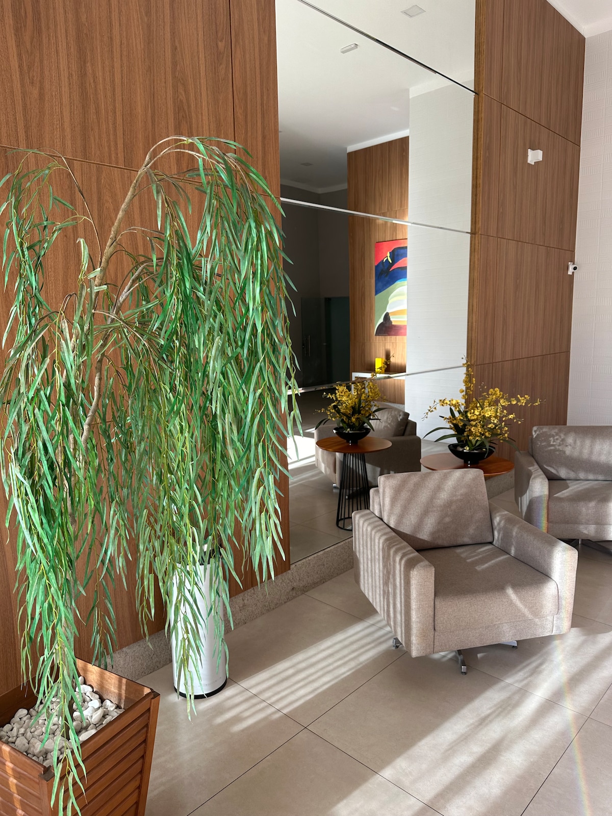 An inviting entrance area features two modern armchairs positioned beside small tables. Decorative plants add a touch of greenery, while a prominent mirror reflects the space. Soft natural light streams in, highlighting the warm wooden paneling and colorful artwork on the wall.