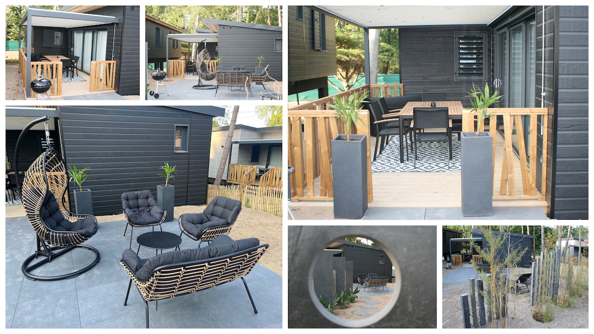 Outdoor seating areas feature a mix of modern furniture, including a circular hanging chair and a lounge set with cushions. A dining table with chairs is positioned on a wooden terrace, surrounded by planters and green foliage. The dark exterior of the cottage provides a contemporary contrast.