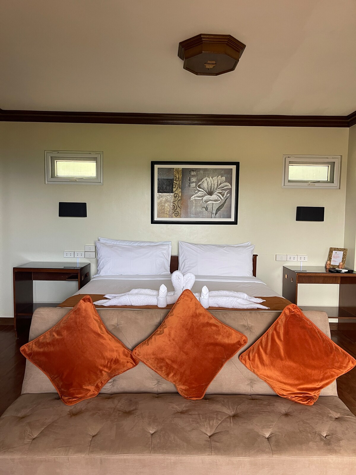 A well-appointed bedroom features two spacious beds flanked by nightstands. A decorative piece of art is displayed above the bed. A cozy seating area with a neutral-colored sofa and orange accent pillows is present in front of the beds.