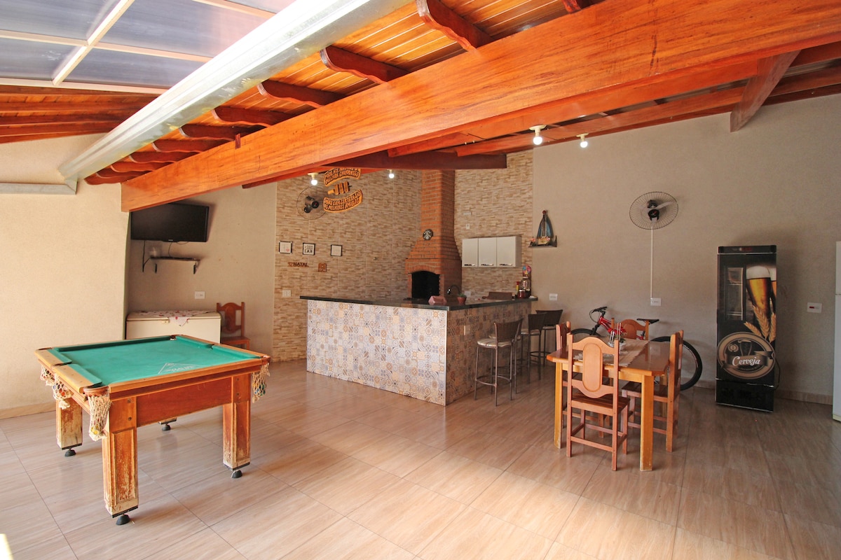 A spacious indoor area features a billiard table, a dining set, and a counter with bar stools. A fan hangs from the ceiling, and a flat-screen television is mounted on a wall. Natural light enters through a skylight, enhancing the open atmosphere.