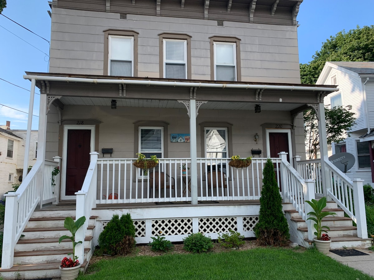 The exterior of a two-story apartment building is displayed, featuring a welcoming porch with white railings and potted plants on either side. Each entrance is marked by a dark door, and light-colored siding contrasts with the greenery of the front lawn.