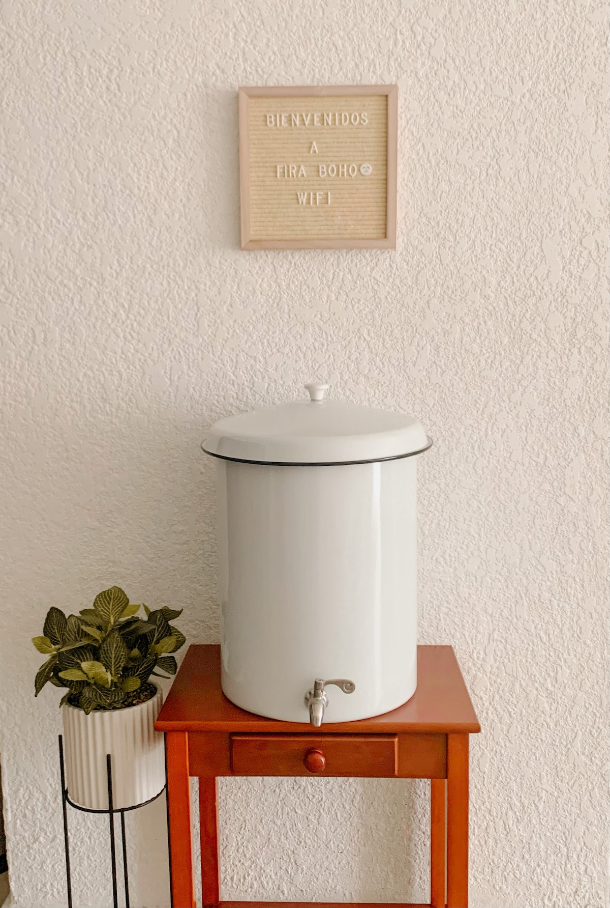 A stylish water dispenser is positioned on a small wooden table with a rich hue. A potted plant adds a touch of greenery, while a welcome sign above features welcoming text. The wall is textured in a soft, light color providing a warm backdrop.