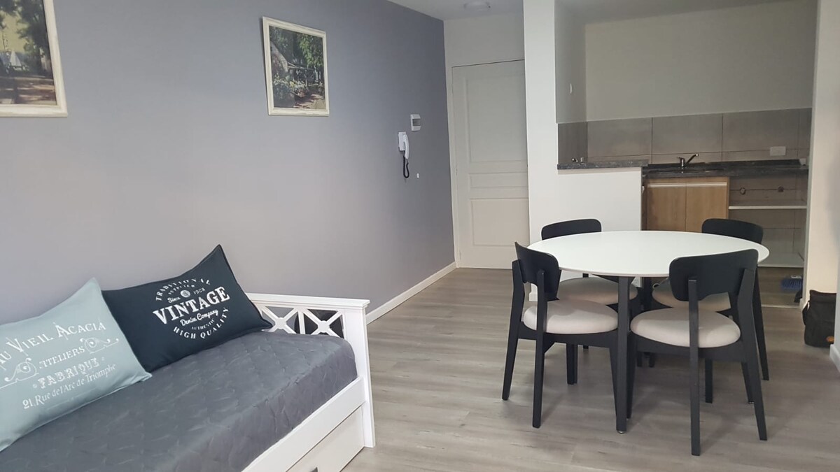 A light-filled living space features a cozy daybed adorned with decorative pillows and a circular dining table surrounded by six chairs. Light-colored walls enhance the overall brightness, while a kitchenette is visible in the background, offering functionality for guests.