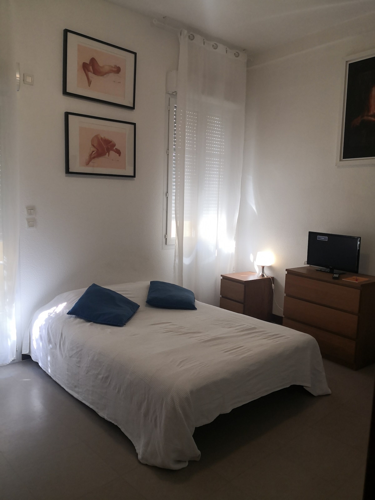 A light-filled sleeping area features a bed with a white coverlet and two blue pillows. A wooden chest of drawers stands nearby, complemented by a small bedside lamp. Art prints adorn the walls, enhancing the space's aesthetic.