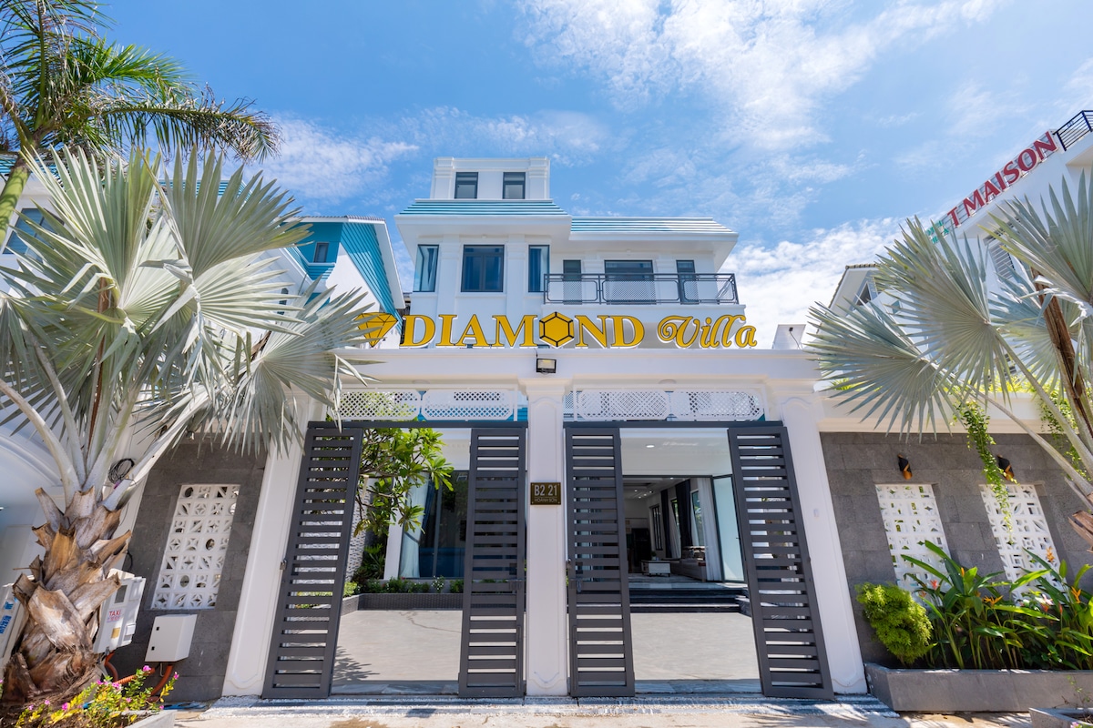 The villa entrance showcases a modern design, framed by palm trees and lush greenery. The name 'Diamond Villa' is prominently displayed above the entrance. The structured facade features a blend of light colors, large windows, and decorative elements, inviting guests into a spacious and welcoming environment.