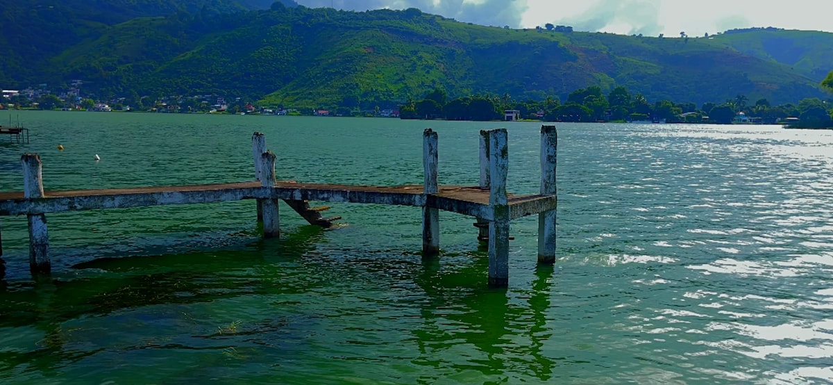 A wooden dock extends over calm waters, with weathered support beams visible. Lush greenery and rolling hills are seen in the background, while the water reflects varying shades of green under a cloudy sky.