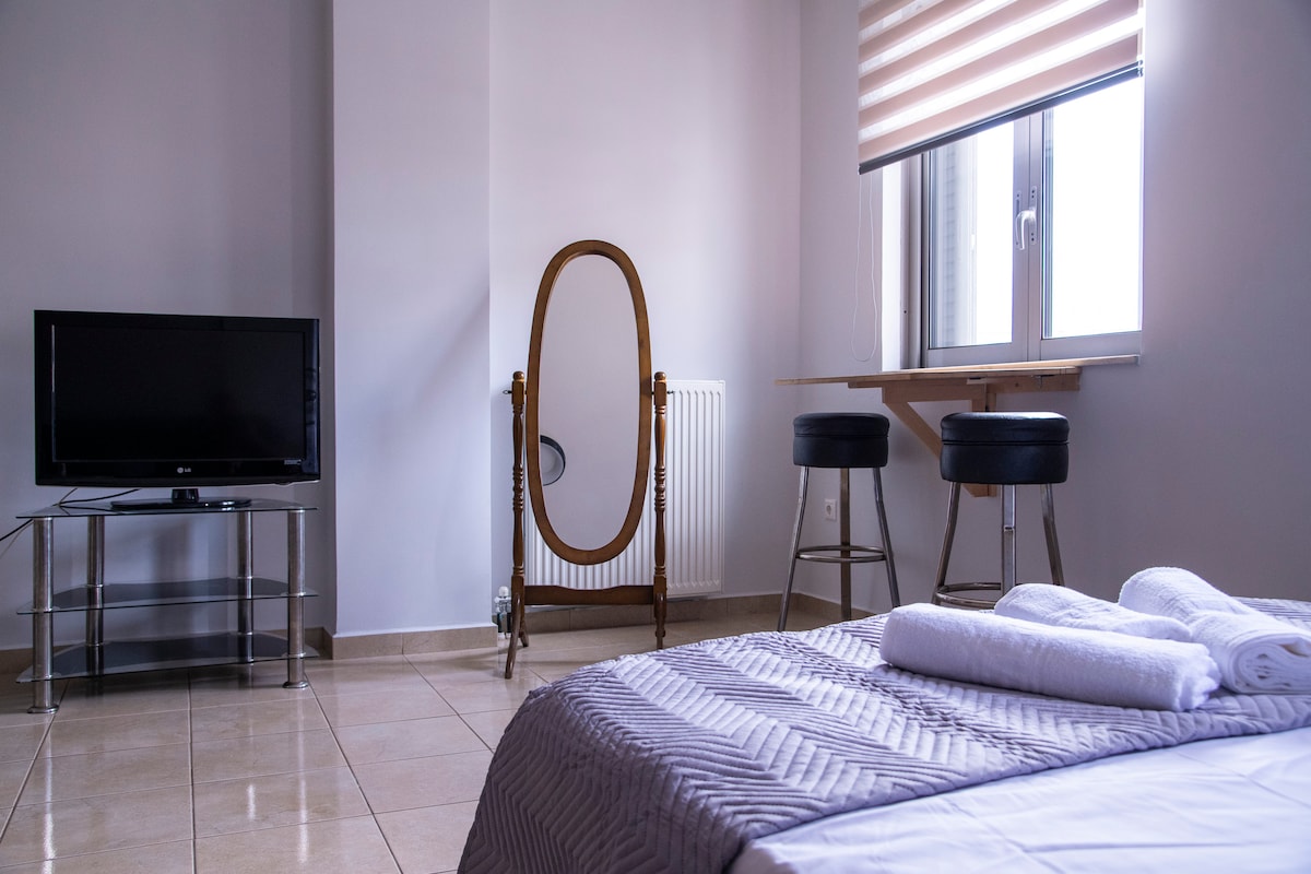A bright, open studio space features a bed with neatly arranged towels. A full-length mirror stands next to a small table with two bar stools. A television is positioned on a glass stand, and a window offers natural light.