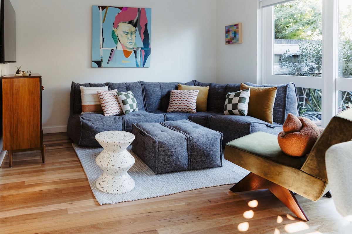 A spacious living area showcases a large sectional sofa adorned with colorful cushions. A mid-century modern accent chair provides additional seating. A light-toned rug lies beneath a sculptural side table. Natural light floods the room through expansive windows, highlighting the vibrant artwork on the wall.