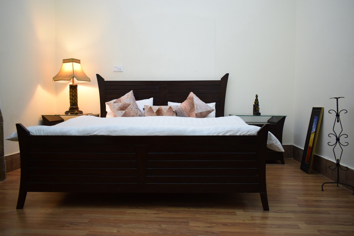 A spacious bed is centered in the room, surrounded by decorative pillows. A bedside lamp provides soft lighting, while a modern side table holds additional decor. The wooden floor adds warmth to the space, complemented by a decorative floor stand and framed artwork against the wall.