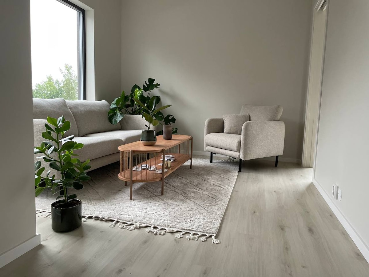 A living area features a light-colored sofa and an armchair alongside a central coffee table. Green plants are positioned in pots, adding a touch of nature to the space. A large window allows natural light to illuminate the room, enhancing the contemporary design.