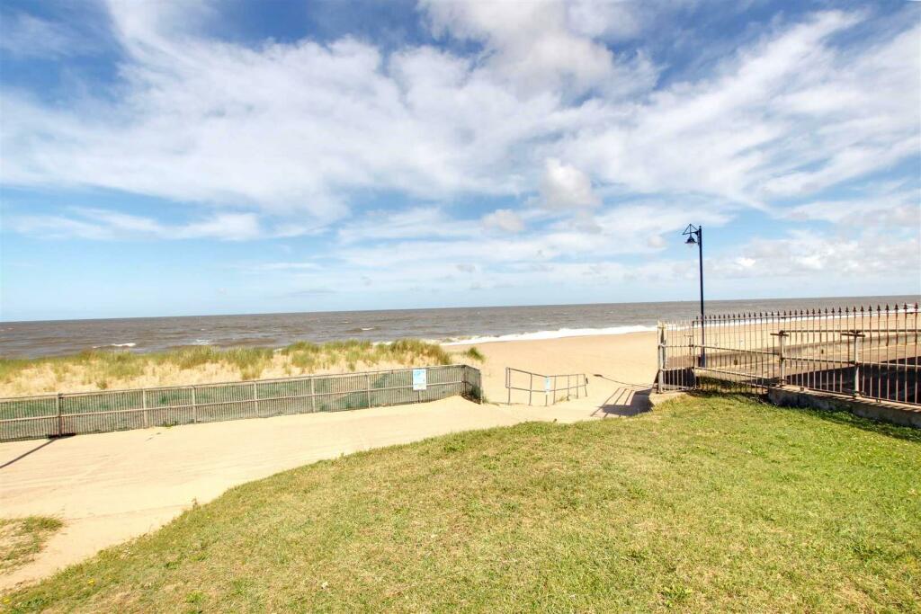 The image captures a sandy beach extending towards the water, framed by green grass. A pathway leads from the grassy area to the beach, with a low fence and a single lamppost visible. The sky is partially cloudy, creating a bright coastal atmosphere.