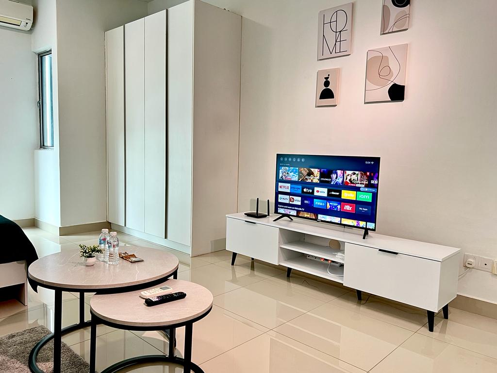 A modern living area features a sleek white TV cabinet with a 40-inch smart TV, displaying various streaming options. Two small round coffee tables are positioned nearby. A built-in wardrobe, with a minimalist design, stands in the background, accompanied by wall-mounted artwork. Natural light filters through the window.