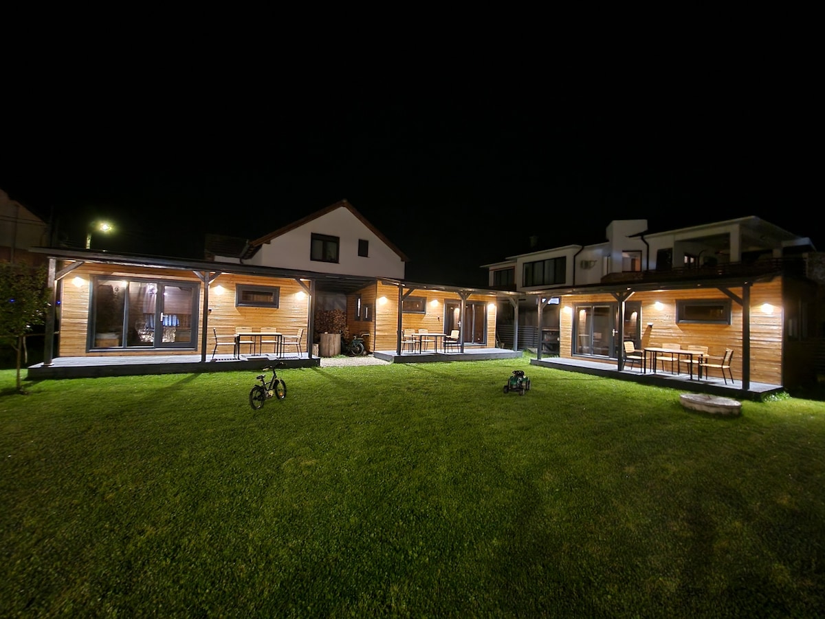 A spacious outdoor area is illuminated by soft lighting, showcasing a well-kept lawn. Wooden terraces host tables and chairs, providing dining options under the stars. Bicycles are parked on the grass, indicating a family-friendly environment.