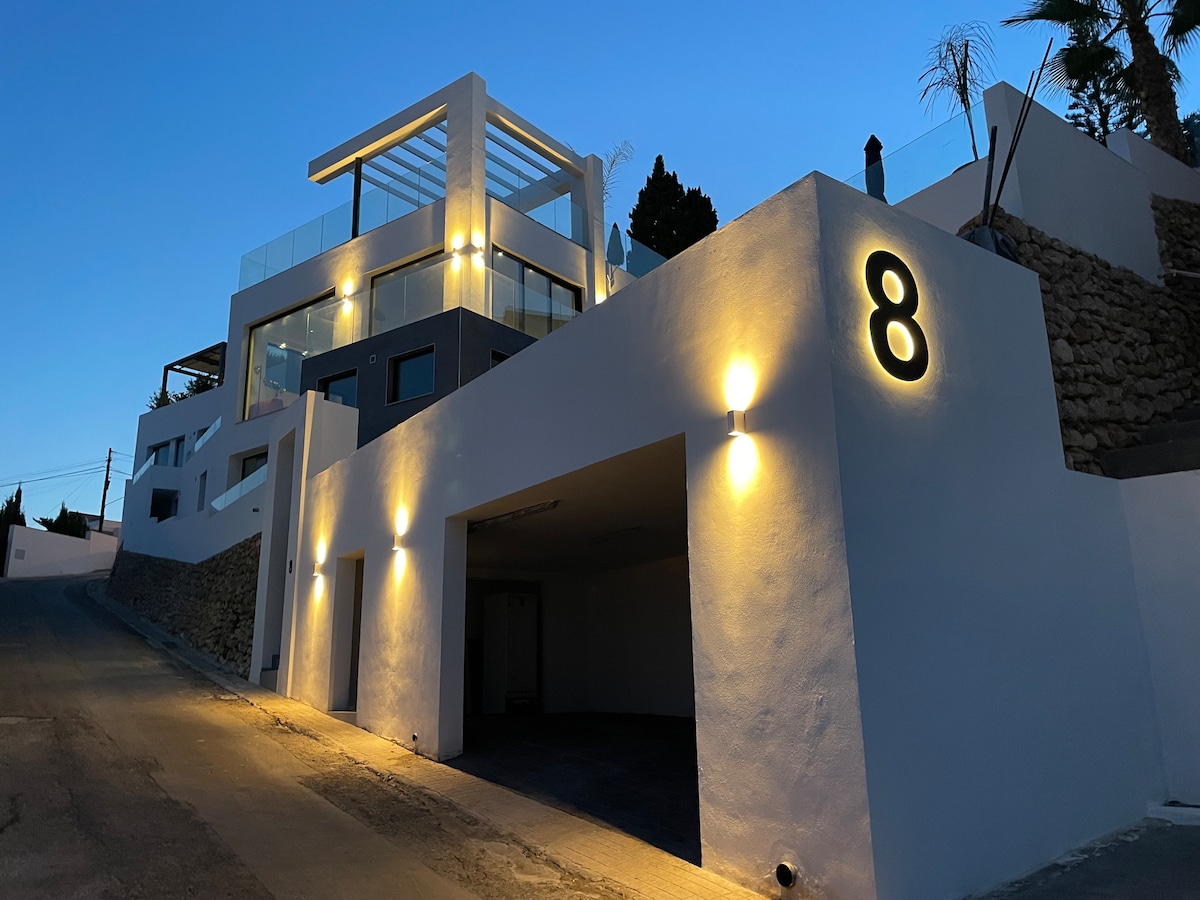 An elegant multi-level building is shown, featuring a modern architectural design. The entrance is illuminated with warm lights, highlighting the large garage area below. A prominent house number '8' is displayed on the exterior wall, with a clear sky in the background.
