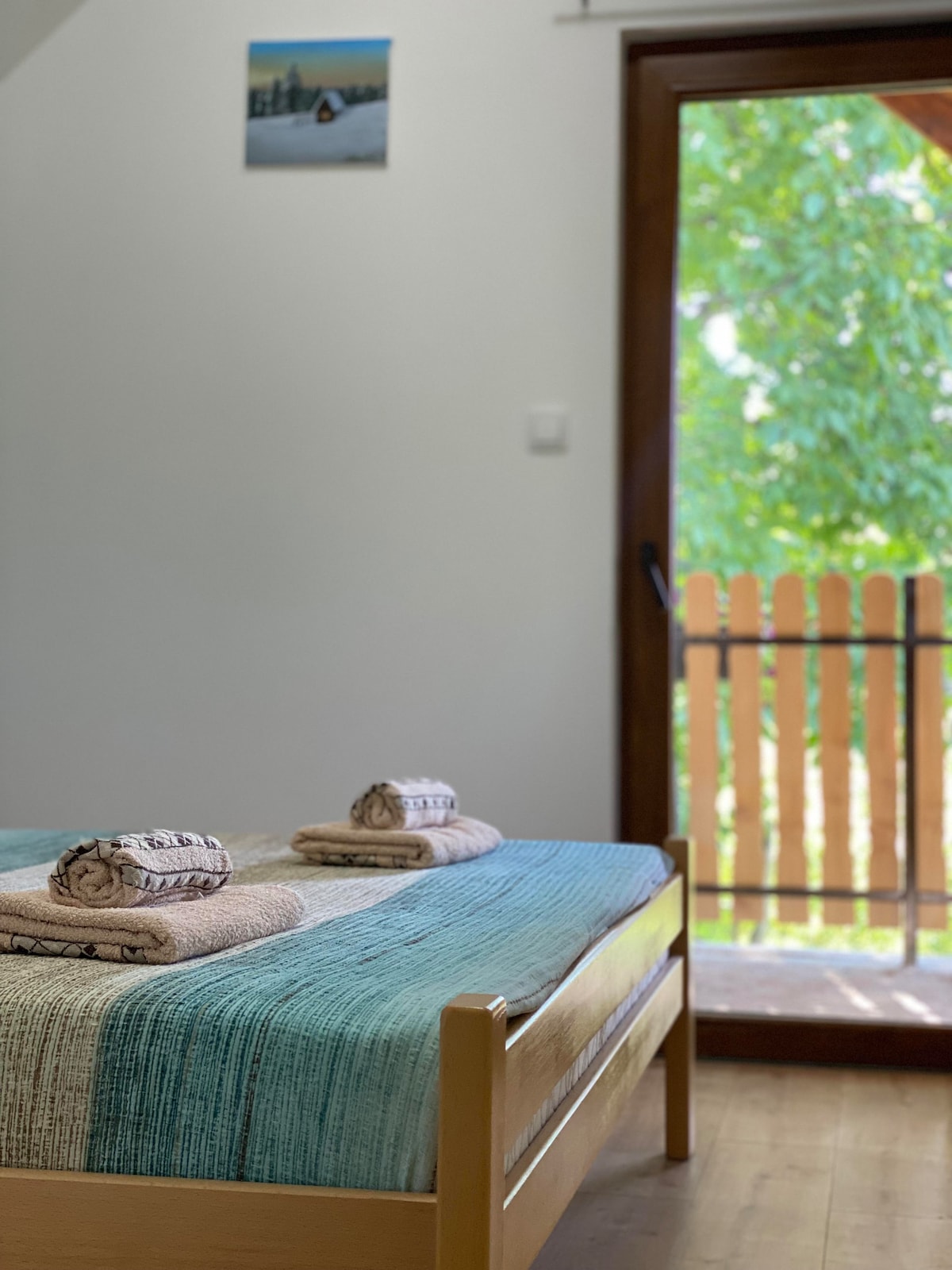 A spacious bed is layered with a textured blanket featuring shades of blue. Two neatly rolled towels are placed on the bed. A large window with a wooden frame overlooks a sunny outdoor area, enhancing the room's light and airy feel.