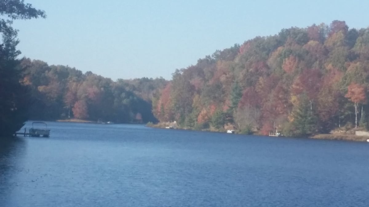 A serene lake scene is captured, bordered by trees displaying autumn foliage in vibrant hues of red, orange, and yellow. The calm water reflects the colorful landscape, while small boats are visible along the shoreline, enhancing the tranquil atmosphere.