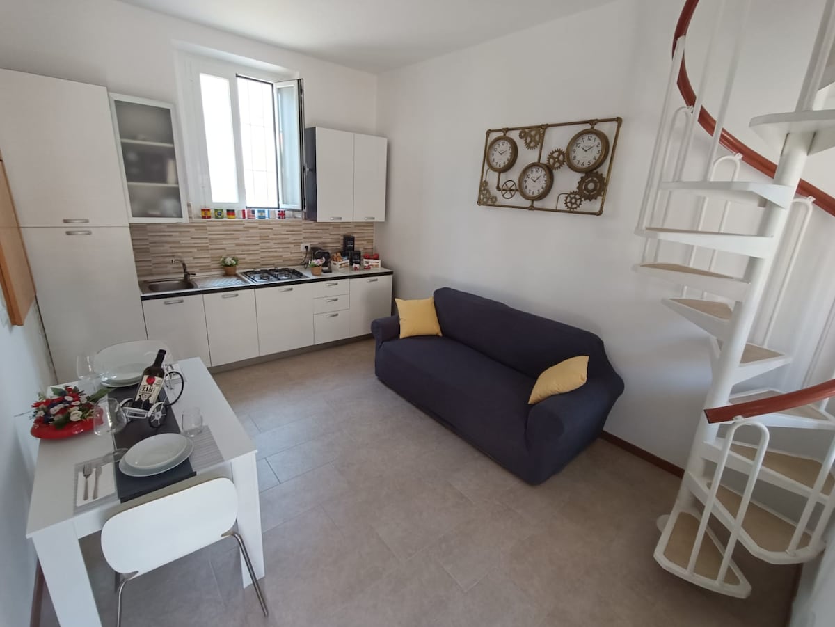 A bright living area features a spacious navy sofa with yellow accent cushions, complemented by a compact dining table set for two. The kitchen area displays modern cabinetry and a tile backsplash, while a spiral staircase leads to the upper level. Natural light enters through a large window.