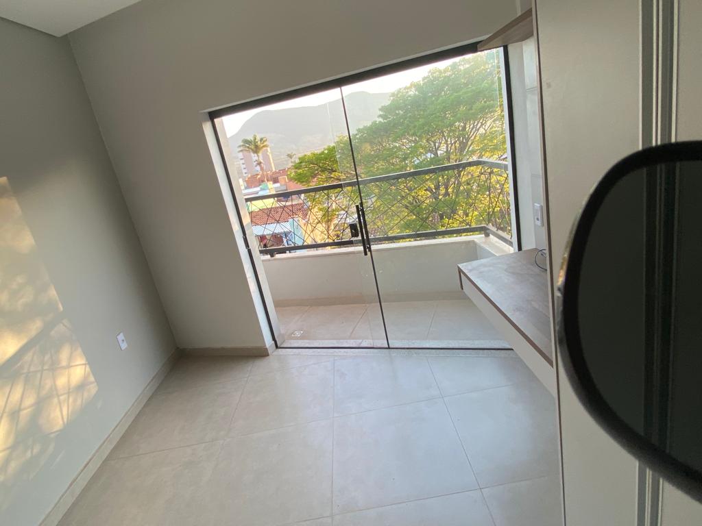 A bright room features large glass doors that open to a balcony, offering views of the surrounding greenery and cityscape. Natural light fills the space, highlighting the soft gray walls and tiled floor, while a small seating area is positioned near the door.