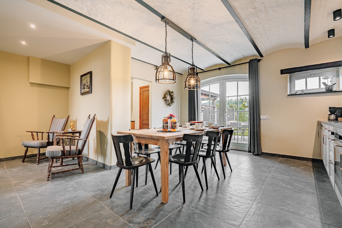 A spacious dining area features a solid wooden table surrounded by black chairs, inviting group meals. Two stylish pendant lights hang above, illuminating the space. A cozy seating arrangement is observed to the left, with large glass doors offering a view of the garden.