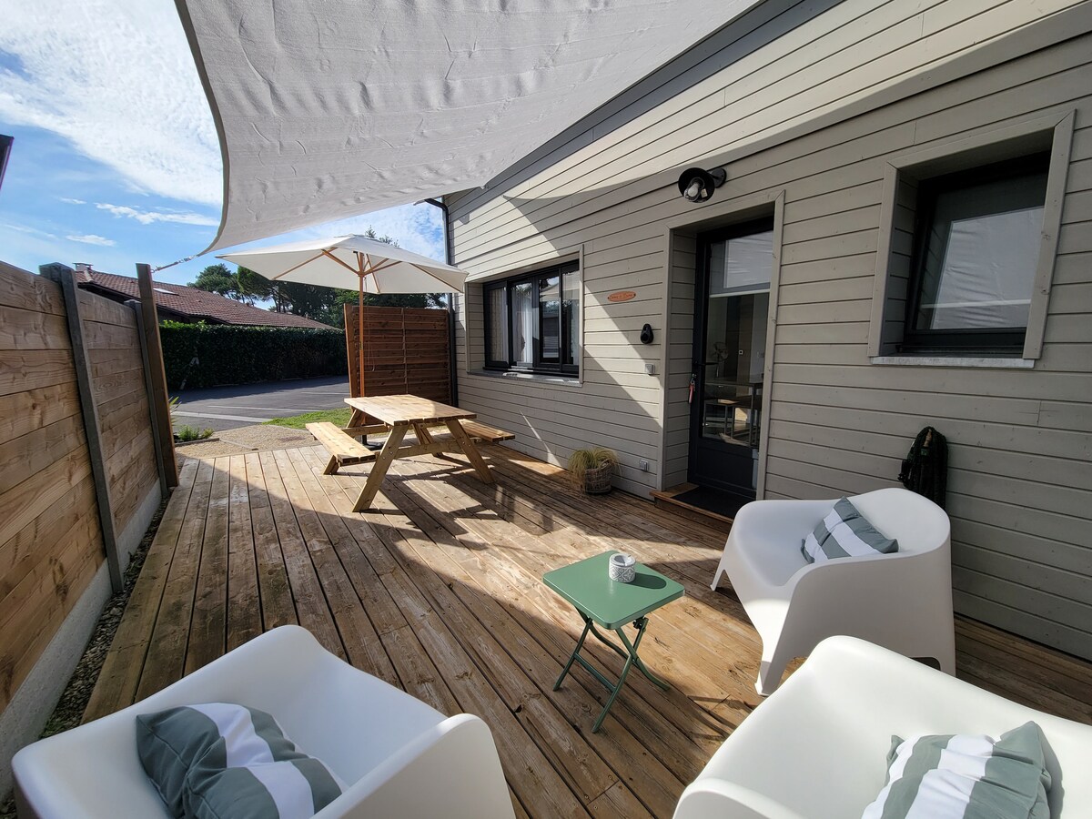 A wooden terrace is featured, with a picnic table and two white armchairs. A small green side table is positioned between the chairs. A large umbrella provides shade, while sunlight illuminates the space, enhancing the inviting outdoor area.