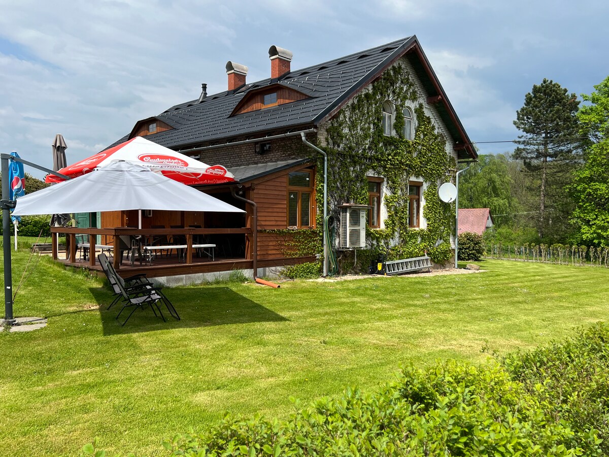 A charming home features a green facade partially covered in climbing vines. A covered outdoor terrace is situated in front, equipped with chairs and a large umbrella for shade. Lush grass surrounds the property, providing an inviting outdoor space.