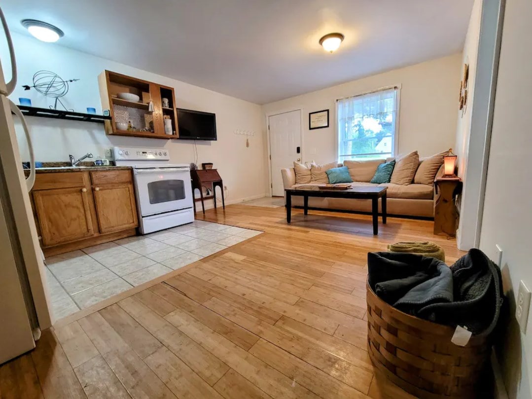 A warm living space with hardwood flooring is complemented by a cozy sofa and coffee table. A fully equipped kitchen area is visible, featuring wooden cabinets and appliances. Natural light filters in through a window, enhancing the inviting atmosphere of the room.