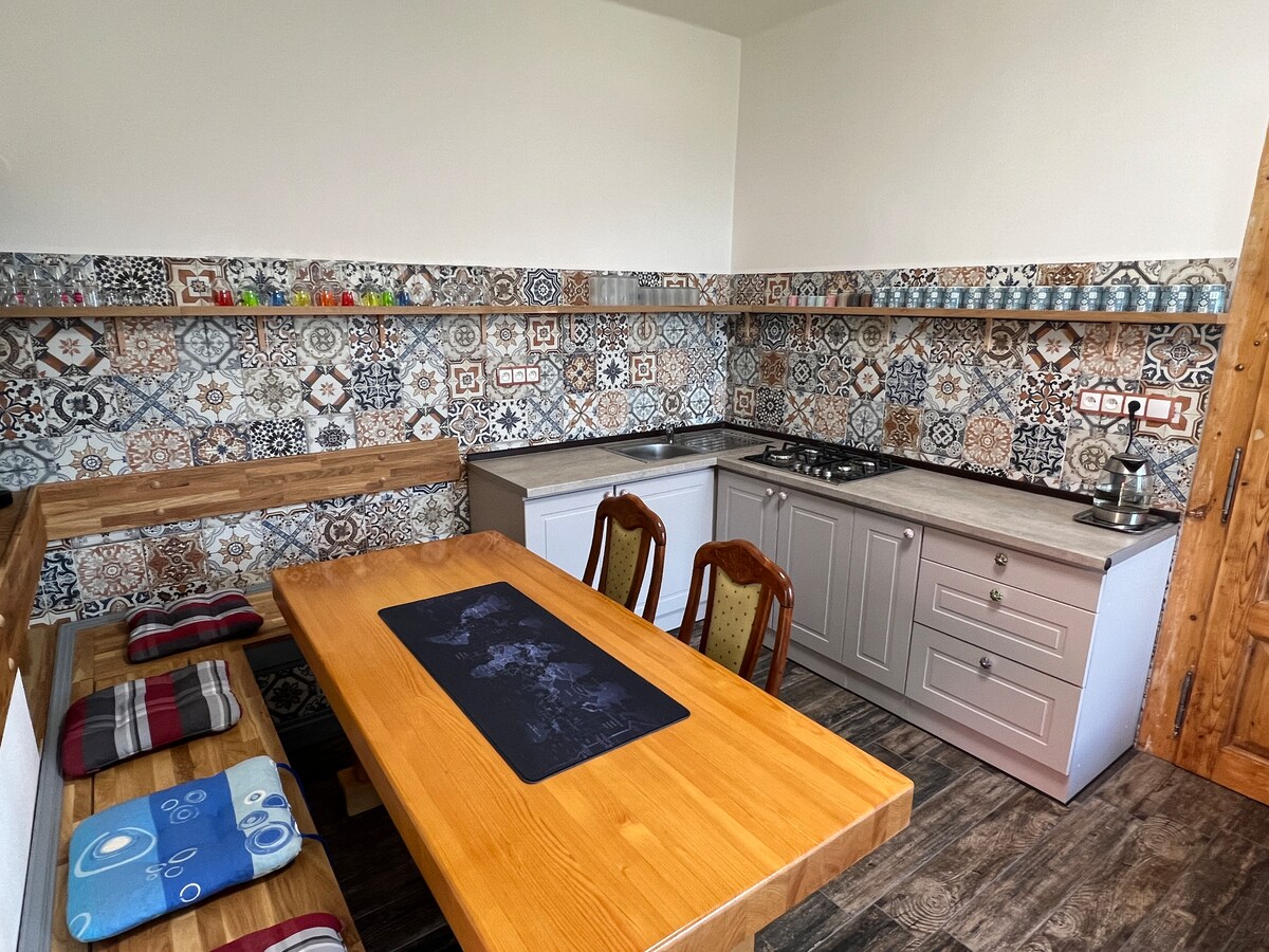 A functional kitchen area features patterned tiles that provide visual interest. The wooden dining table is accompanied by several chairs, while the kitchen counter is equipped with essential appliances and utensils, inviting culinary creativity.