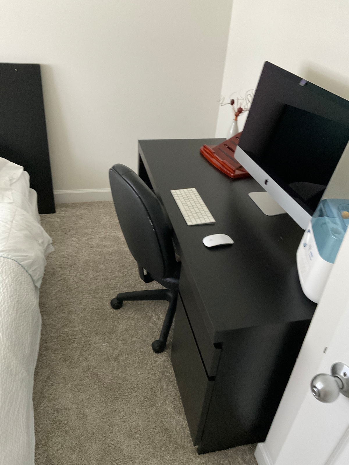 A well-organized workspace is shown, featuring a sleek black desk with a computer monitor, keyboard, and mouse. A comfortable office chair is positioned nearby. An additional item on the desk is a small appliance, while a cozy bed with white linens is visible in the background.