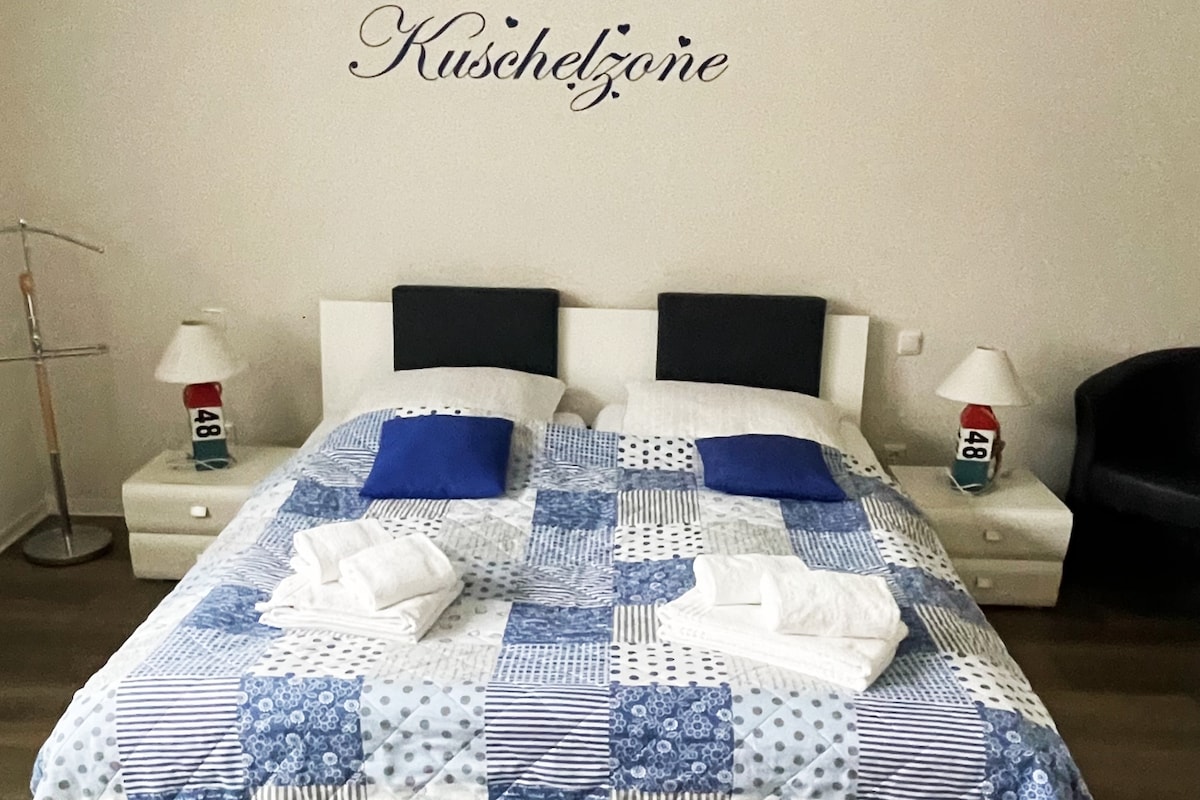 A comfortable sleeping area features a spacious bed with a patchwork quilt laid across it. Two blue cushions are placed on the pillows, and bedside tables with lamps are positioned at either side. A cozy armchair is visible in the corner, enhancing the room's inviting atmosphere.