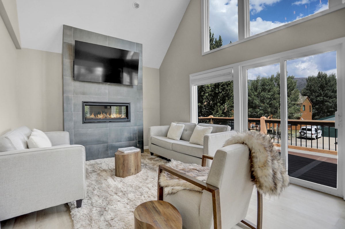 A spacious living area features large windows framing mountain views, allowing natural light to fill the room. A cozy fireplace rests beneath a flat-screen TV. Comfortable sofas are arranged around a central wooden table, complemented by a soft area rug underfoot.