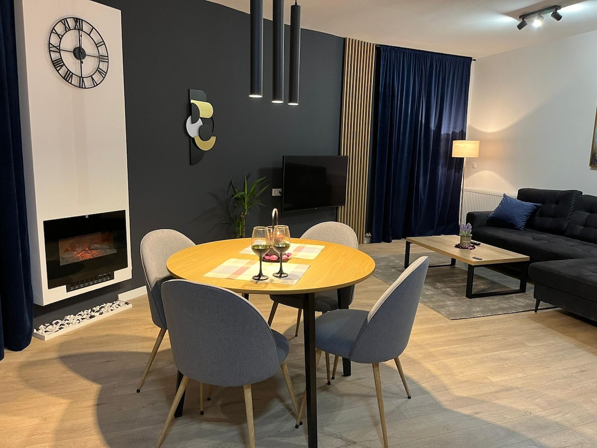 A modern dining area features a round wooden table surrounded by four upholstered chairs. A stylish clock is mounted on the wall, and a sleek electric fireplace adds warmth. A comfortable sectional sofa and a floor lamp create an inviting space in the background.