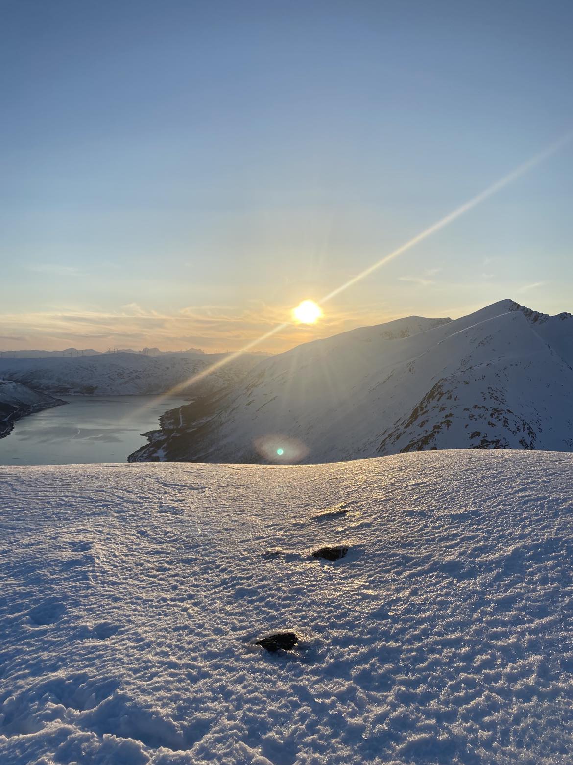A stunning winter landscape is captured, revealing a snow-covered terrain illuminated by the setting sun. The sun's rays create a warm glow, while distant mountains and a winding water body are visible, offering a serene and picturesque view.