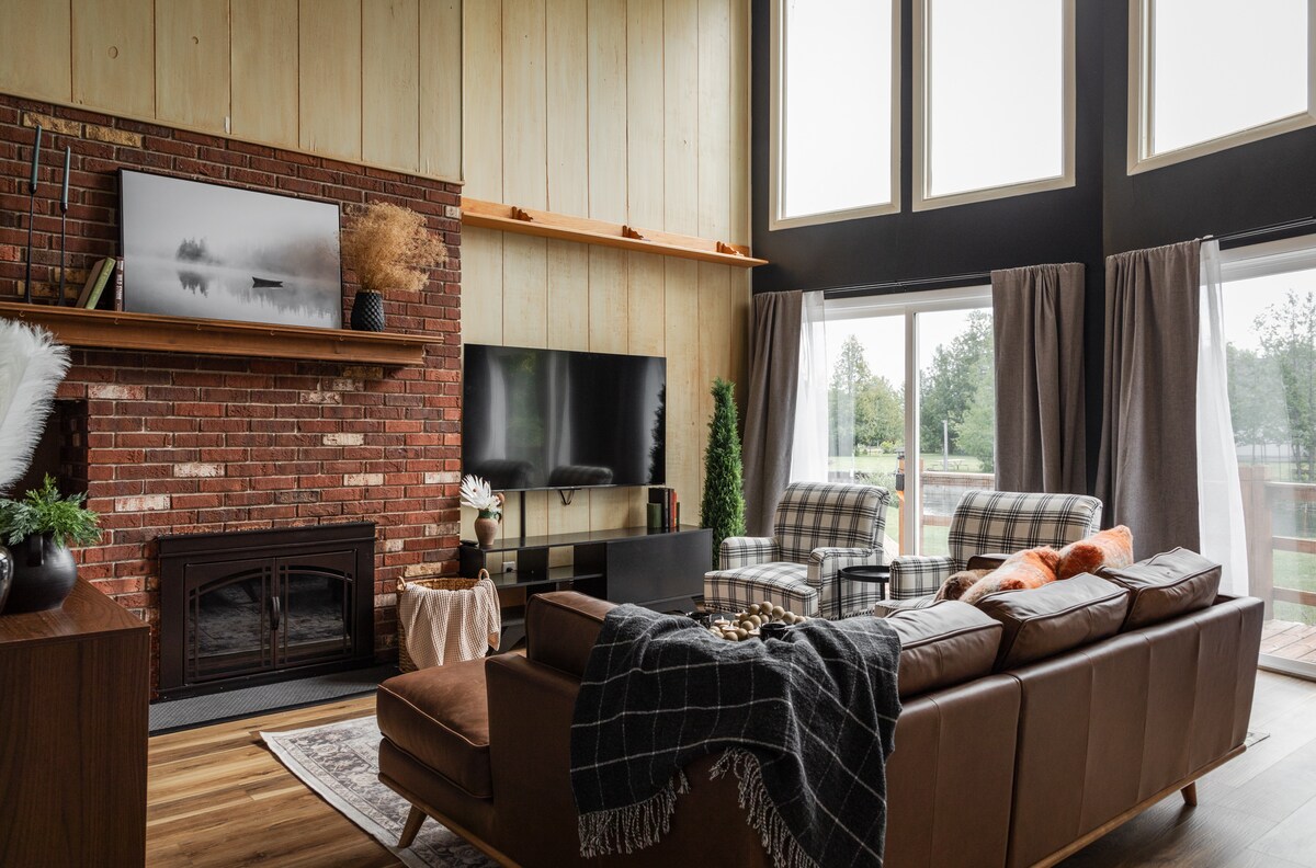 A spacious living area features large windows, allowing natural light to fill the room. The brick fireplace is complemented by a modern television. A comfortable brown sofa is adorned with a patterned throw, and cozy chairs provide additional seating options.