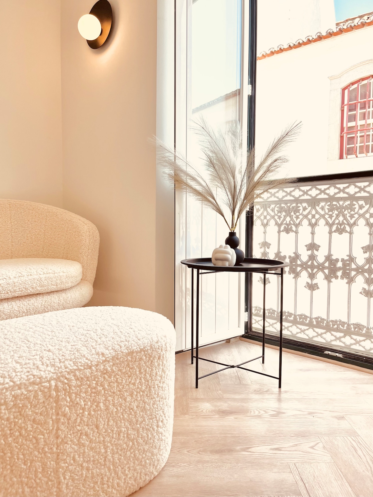 A cozy corner of the living area features a soft, textured sofa paired with a round footstool. A minimalist side table displays a vase with decorative pampas grass, positioned beside large glass doors that open to a traditional balcony railing.