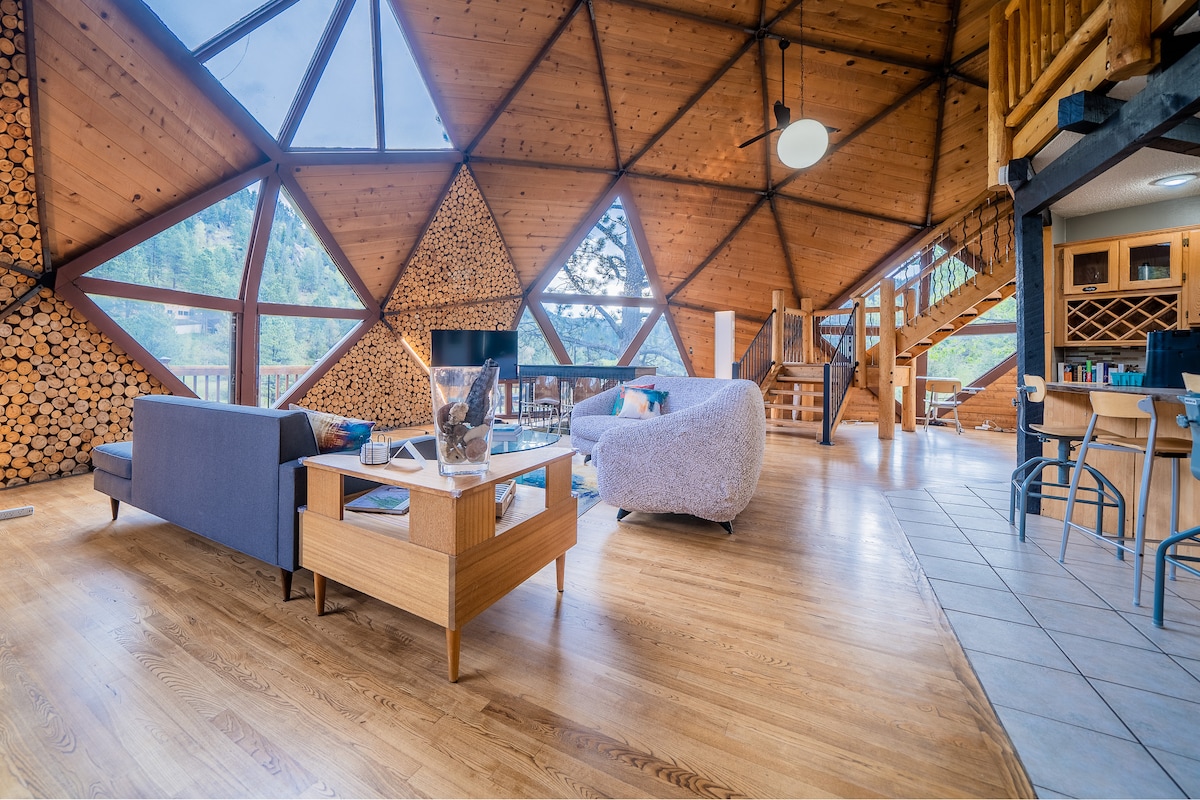 The interior of the Dome showcases a high, curved ceiling with large triangular windows allowing natural light to flood the space. A cozy seating area is furnished with modern couches and a wooden coffee table. A staircase leads to an upper level, while wood accents and stacked logs add warmth.