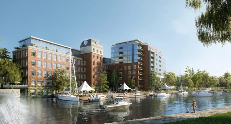 A waterfront view showcases a modern brick building with multiple stories and large windows. A marina with various boats is visible in front, complemented by nearby trees and tents. Clear skies and sunlight illuminate the serene atmosphere of the setting.