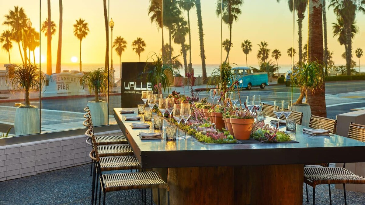 A large outdoor dining table, surrounded by palm trees, is set for a meal with multiple glasses and decorative plants. In the background, the sun sets over the Pacific Ocean, casting a warm glow on the scene. A glimpse of Oceanside Pier is seen in the distance.