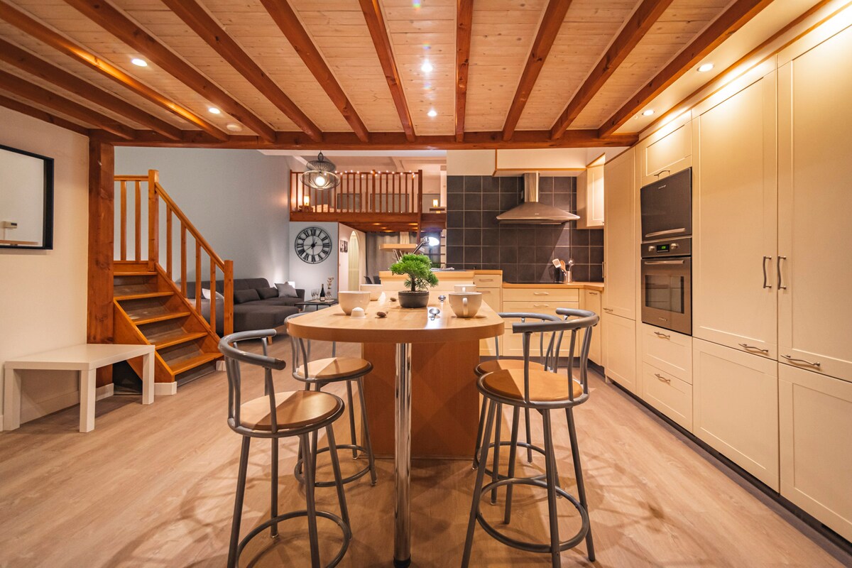 The kitchen area features wooden beams overhead and includes a central round table with four stools. Modern cabinetry in light tones provides ample storage, while high-end appliances, including an oven and stovetop, are integrated seamlessly. Stairs lead to the upper level in the background.