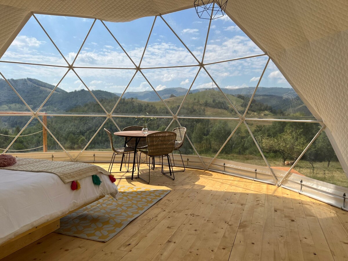A spacious geodesic dome interior features panoramic windows that offer expansive views of the surrounding mountains and landscape. A wooden table and matching chairs sit under a geometrically designed light fixture, with a cozy bed seen in the foreground adorned with colorful decorative elements.
